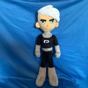 Peut inclure: Un jouet en peluche représentant un personnage de dessin animé avec des cheveux blancs, un t-shirt noir avec un "P" blanc et un pantalon gris. Le personnage a une expression sérieuse avec des yeux verts.