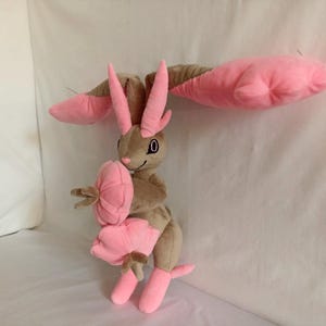 Puede incluir: Un juguete de peluche con forma de conejo con orejas rosas y una flor rosa en su mano. El conejo es marr&oacute;n y tiene un ojo negro.