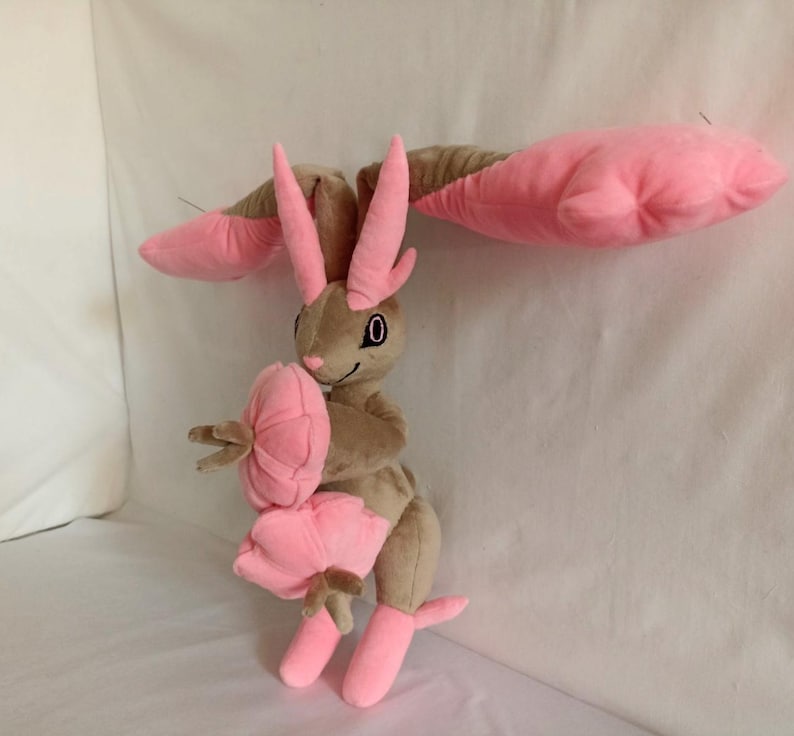 Puede incluir: Un juguete de peluche con forma de conejo con orejas rosas y una flor rosa en su mano. El conejo es marr&oacute;n y tiene un ojo negro.