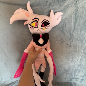 May include: A pink and white plush toy with long arms and a black bow tie. The toy has a detailed face with yellow and red eyes and a toothy grin.