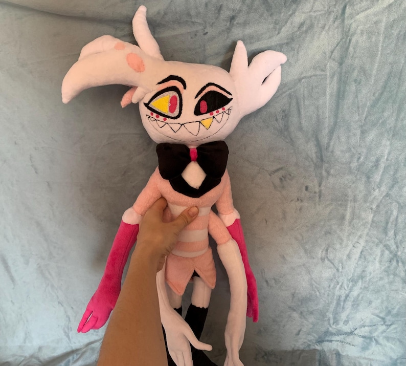 May include: A pink and white plush toy with long arms and a black bow tie. The toy has a detailed face with yellow and red eyes and a toothy grin.