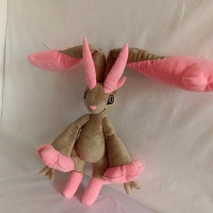 Puede incluir: Un juguete de peluche con forma de conejo con orejas largas de color rosa y pelaje marr&oacute;n. El juguete tiene grandes pies rosas y una cara sonriente con ojos negros.