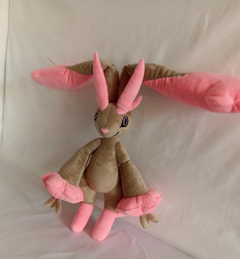 Puede incluir: Un juguete de peluche con forma de conejo con orejas largas de color rosa y pelaje marr&oacute;n. El juguete tiene grandes pies rosas y una cara sonriente con ojos negros.