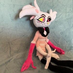 May include: A plush toy of a white and pink character with black horns, yellow eyes, and a pink and white striped dress. The toy has long pink arms and black legs.
