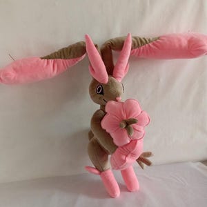 Puede incluir: Un juguete de peluche de un conejo rosa y marr&oacute;n que sostiene una flor rosa. El conejo tiene orejas largas y una cola larga. La flor es un dise&ntilde;o simple con cinco p&eacute;talos.