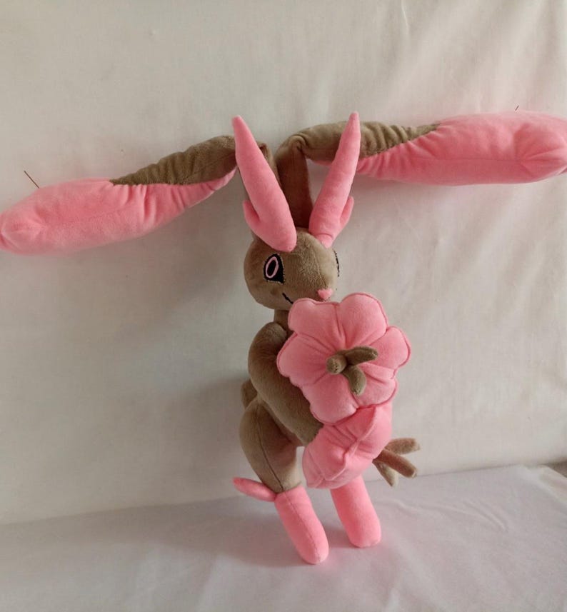 Puede incluir: Un juguete de peluche de un conejo rosa y marr&oacute;n que sostiene una flor rosa. El conejo tiene orejas largas y una cola larga. La flor es un dise&ntilde;o simple con cinco p&eacute;talos.