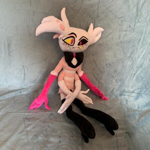 May include: A plush toy of a pink and white character with black horns, yellow eyes, and a pink bow tie. The toy is sitting on a blue fuzzy surface.