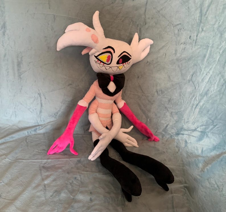 May include: A plush toy of a pink and white character with black horns, yellow eyes, and a pink bow tie. The toy is sitting on a blue fuzzy surface.