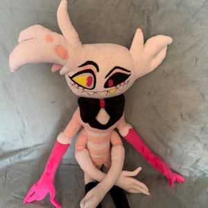 May include: A plush toy of a pink and white character with yellow and red eyes, a black bow tie, and pink arms. The character is sitting on a light gray fuzzy surface.
