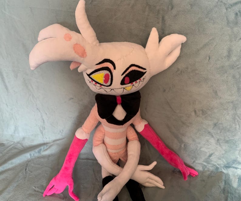 May include: A plush toy of a pink and white character with yellow and red eyes, a black bow tie, and pink arms. The character is sitting on a light gray fuzzy surface.