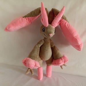 Puede incluir: Un juguete de peluche de conejo con pelaje marr&oacute;n y orejas y patas rosas. El conejo tiene una nariz larga y puntiaguda y ojos grandes y negros.