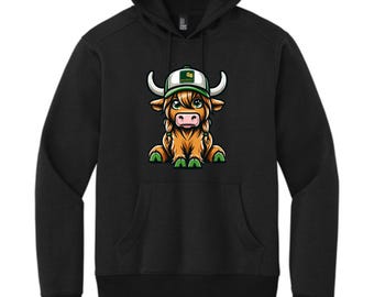 May include: Black hoodie with a cartoon illustration of a Highland cow wearing a green and white cap. The cow has brown fur, green eyes, and green details. The sweatshirt has a front pocket and a hood.