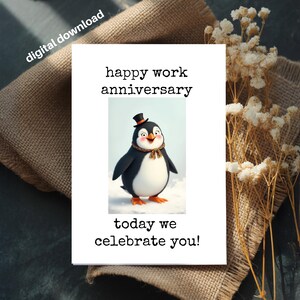 以下が含まれることがあります： 「happy work anniversary」と「today we celebrate you!」の文字が入った白いカードには、シルクハットとスカーフを身につけた漫画のペンギンが描かれています。カードは麻布の表面にあり、ドライフラワーが添えられています。