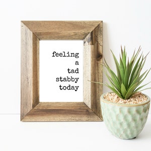 May include: A framed print with the text "feeling a tad stabby today" in a typewriter font. The print is in a wooden frame and is sitting on a white surface next to a potted succulent.