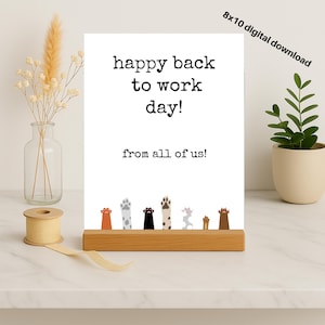Peut inclure: Une carte blanche avec le texte "happy back to work day!" et "from all of us!" est affichée sur un support en bois. La carte présente des empreintes de pattes en bas. Un vase avec des fleurs séchées et une petite plante en pot sont de chaque côté.