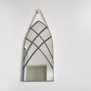 May include: A silver arched mirror with a decorative geometric pattern of intersecting lines. The mirror is hanging on a white wall.