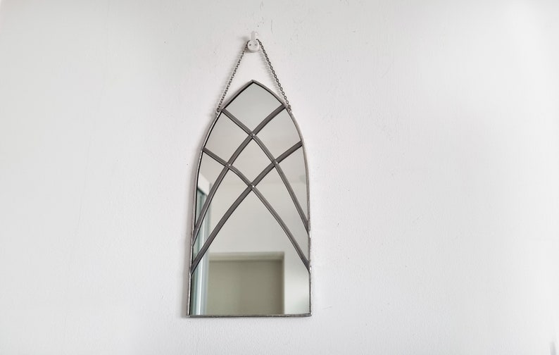 May include: A silver arched mirror with a decorative geometric pattern of intersecting lines. The mirror is hanging on a white wall.