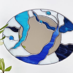Op de afbeelding: Een spiegel van gekleurd glas met een abstract ontwerp in blauw, wit en zwart. De spiegel is ovaalvormig en heeft een zilveren lijst.