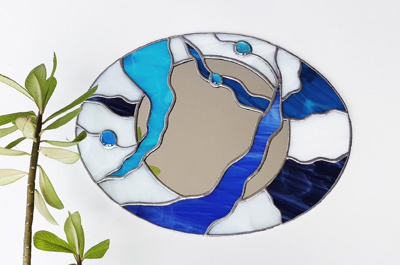 Op de afbeelding: Een spiegel van gekleurd glas met een abstract ontwerp in blauw, wit en zwart. De spiegel is ovaalvormig en heeft een zilveren lijst.