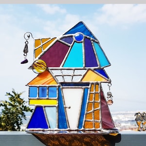 以下が含まれることがあります： 青、紫、オレンジ、黄色の色合いで幾何学模様が施されたステンドグラスの家。家は白いドアがあり、ぶら下がっているチャームで飾られています。