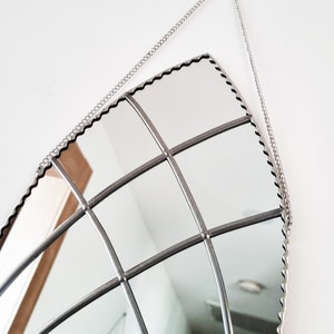 May include: A silver-toned, decorative mirror with a scalloped edge and a grid pattern. The mirror is hanging from a silver chain.