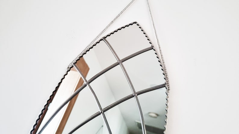 May include: A silver-toned, decorative mirror with a scalloped edge and a grid pattern. The mirror is hanging from a silver chain.