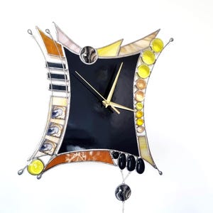 May include: A wall clock with a central black square face and gold hands. The frame is constructed from stained glass pieces in shades of yellow, amber, and black, featuring a pendulum with a circular patterned disc hanging from the bottom.