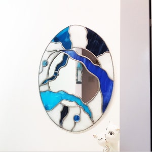 Op de afbeelding: Een ronde spiegel van gekleurd glas met een abstract ontwerp in blauw, wit en zwart. De spiegel is ingelijst in zilver.