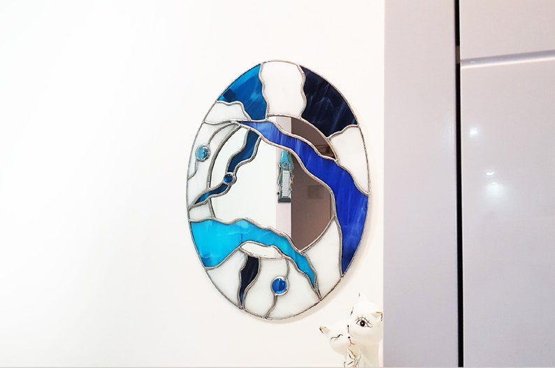 Op de afbeelding: Een ronde spiegel van gekleurd glas met een abstract ontwerp in blauw, wit en zwart. De spiegel is ingelijst in zilver.