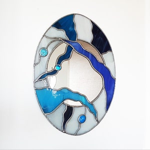 Op de afbeelding: Ovale spiegel met een glas-in-lood frame met een abstract ontwerp in blauw, wit en zwart. Het frame is gemaakt van kleine stukjes glas die met lood aan elkaar zijn verbonden.