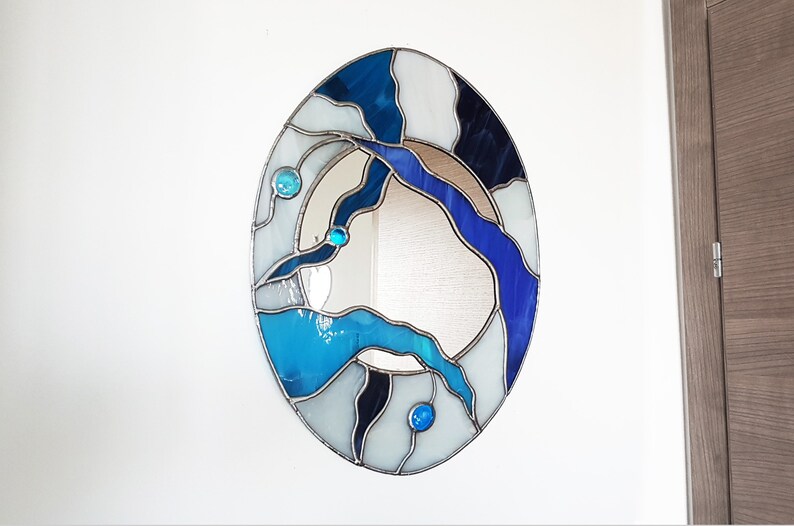 Op de afbeelding: Ovale spiegel met een glas-in-lood frame met een abstract ontwerp in blauw, wit en zwart. Het frame is gemaakt van kleine stukjes glas die met lood aan elkaar zijn verbonden.