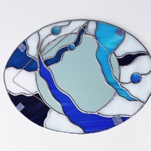 Op de afbeelding: Ovale spiegel van gekleurd glas met een abstract ontwerp in blauw, wit en zwart. De spiegel is omgeven door een zilveren lijst.