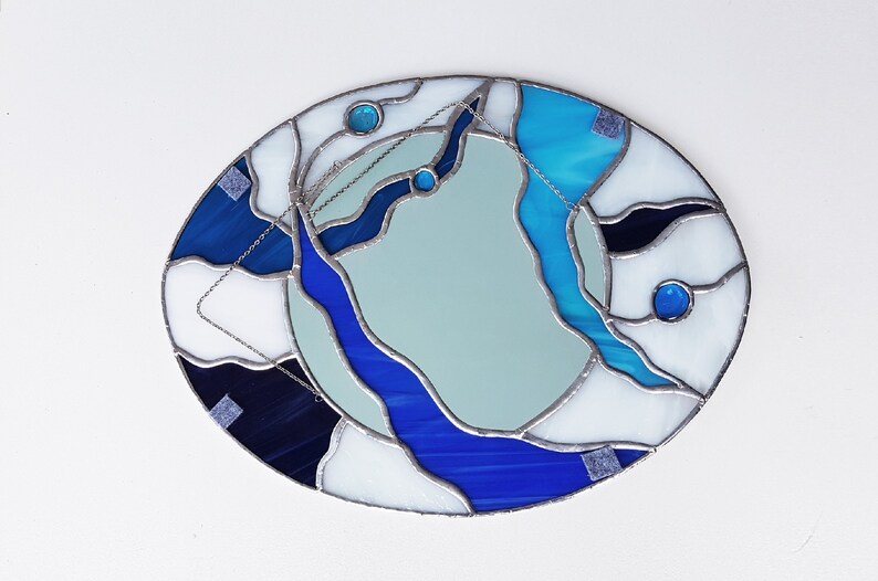 Op de afbeelding: Ovale spiegel van gekleurd glas met een abstract ontwerp in blauw, wit en zwart. De spiegel is omgeven door een zilveren lijst.