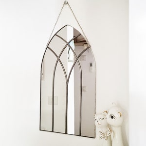 May include: A silver arched mirror with a decorative metal frame hangs on a white wall. The mirror is reflecting a white wall and a doorway. Two white ceramic figurines are in the bottom right corner of the image.