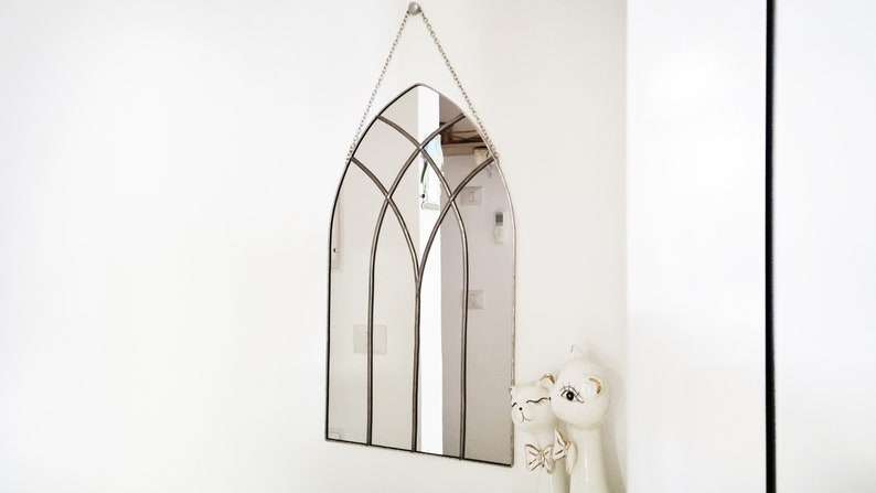 May include: A silver arched mirror with a decorative metal frame hangs on a white wall. The mirror is reflecting a white wall and a doorway. Two white ceramic figurines are in the bottom right corner of the image.