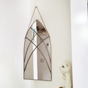May include: A silver arched mirror with a decorative beaded border and a geometric design in the center. The mirror is hanging on a white wall.