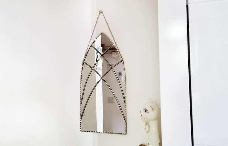 May include: A silver arched mirror with a decorative beaded border and a geometric design in the center. The mirror is hanging on a white wall.