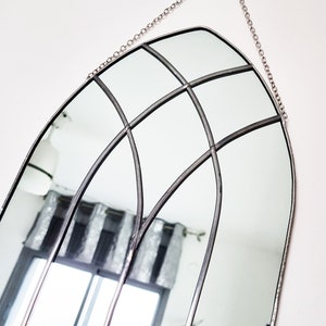 May include: A silver arched mirror with a decorative metal frame and a chain for hanging. The mirror is reflecting a room with a window and curtains.