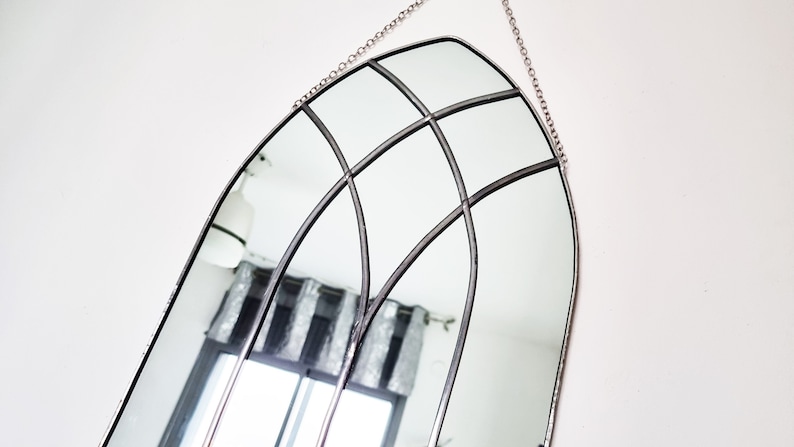 May include: A silver arched mirror with a decorative metal frame and a chain for hanging. The mirror is reflecting a room with a window and curtains.