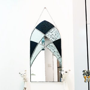 May include: A black and white stained glass mirror with a gothic arch design. The mirror is hanging on a white wall.