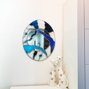 Op de afbeelding: Een ronde spiegel van gekleurd glas met blauwe en witte glasstukken. De spiegel hangt aan een witte muur met twee witte keramische kattenbeeldjes ervoor.