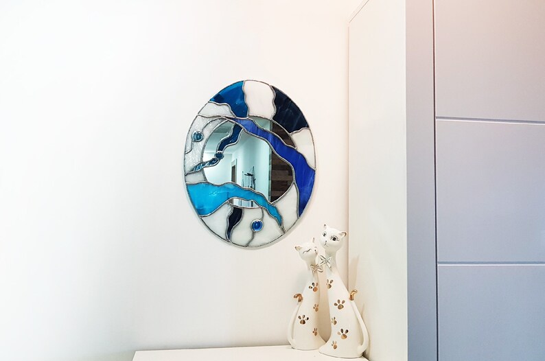 Op de afbeelding: Een ronde spiegel van gekleurd glas met blauwe en witte glasstukken. De spiegel hangt aan een witte muur met twee witte keramische kattenbeeldjes ervoor.