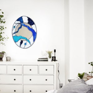 Op de afbeelding: Een witte commode met zes laden en zwarte knoppen. Een ronde spiegel van gekleurd glas met blauwe, witte en zwarte abstracte ontwerpen hangt boven de commode. Een kleine plant in een pot staat op de commode naast een zwarte lamp.