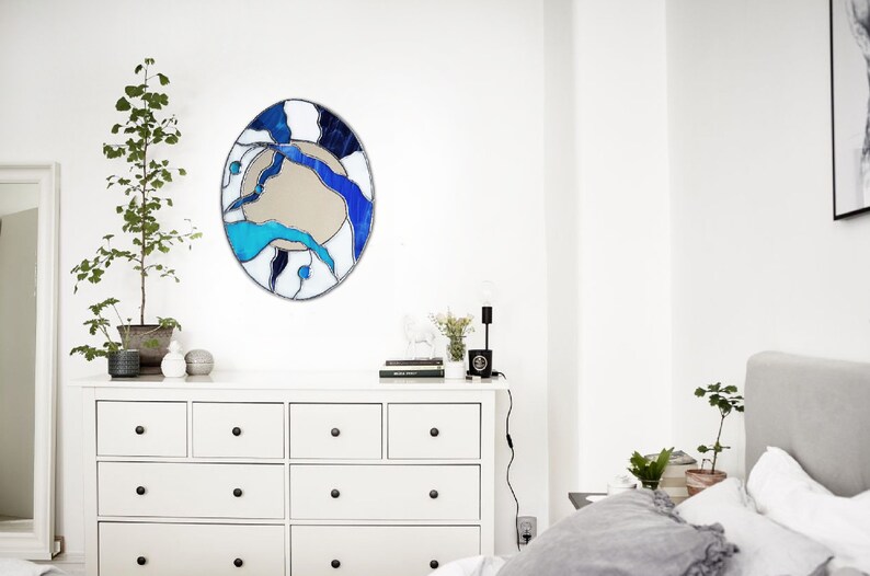 Op de afbeelding: Een witte commode met zes laden en zwarte knoppen. Een ronde spiegel van gekleurd glas met blauwe, witte en zwarte abstracte ontwerpen hangt boven de commode. Een kleine plant in een pot staat op de commode naast een zwarte lamp.