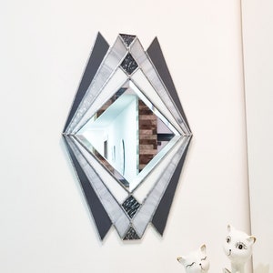 Op de afbeelding: Een geometrische spiegel van gekleurd glas in de vorm van een diamant met een zilver- en grijs kleurenschema. De spiegel is gemonteerd op een witte muur.