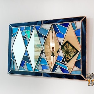 Pode incluir: Um espelho retangular de vidro colorido com um design geométrico que apresenta três formas de diamante. Os diamantes são feitos de peças de vidro azul, branco e dourado. O espelho é emoldurado em azul escuro.