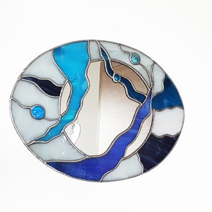 Op de afbeelding: Een ronde spiegel van gekleurd glas met een abstract ontwerp in blauw, wit en zwart. De spiegel heeft een zilveren lijst en is omgeven door een witte muur.