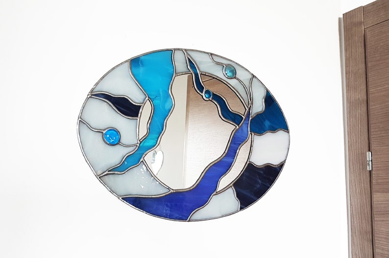 Op de afbeelding: Een ronde spiegel van gekleurd glas met een abstract ontwerp in blauw, wit en zwart. De spiegel heeft een zilveren lijst en is omgeven door een witte muur.