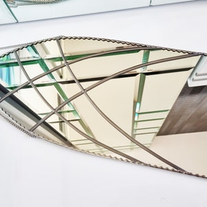 May include: A decorative, silver-toned, beveled, oval-shaped mirror with a scalloped edge. The mirror is hanging from a silver chain and reflects a view of a room with a staircase.