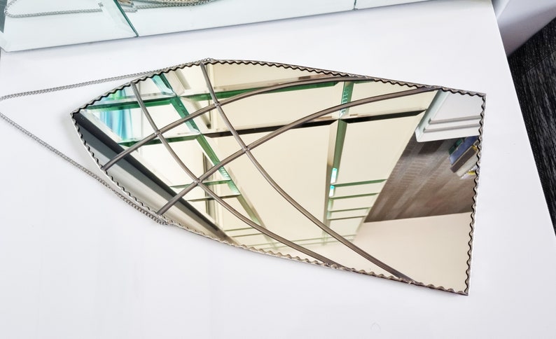 May include: A decorative, silver-toned, beveled, oval-shaped mirror with a scalloped edge. The mirror is hanging from a silver chain and reflects a view of a room with a staircase.